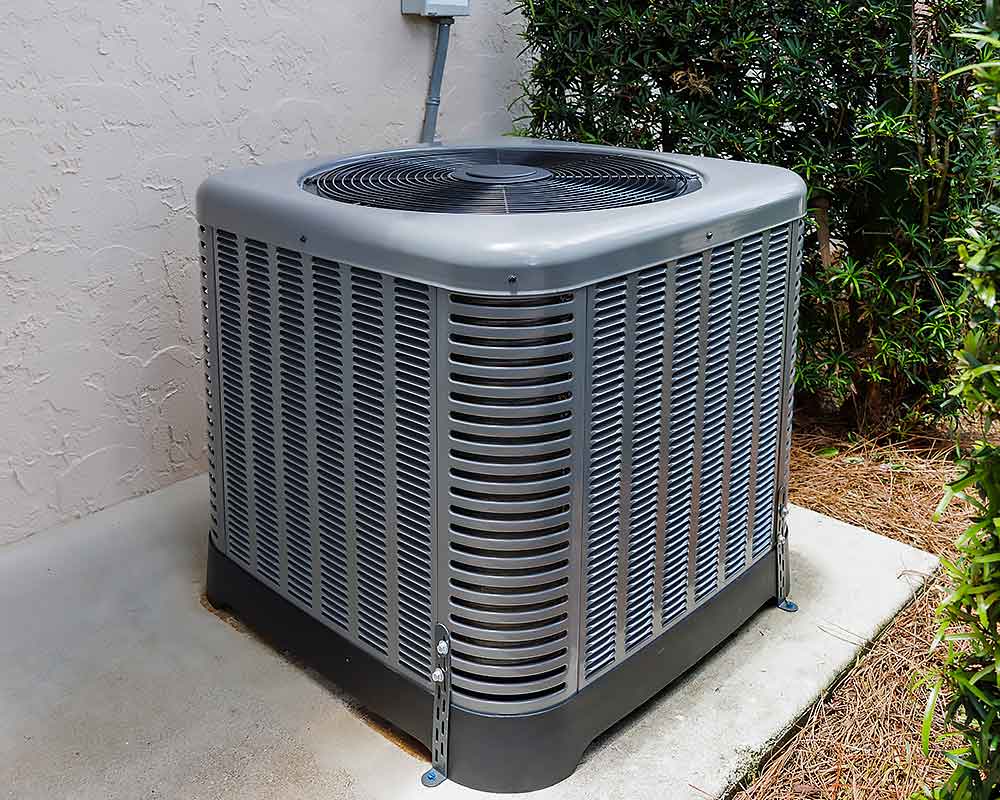 A gray outdoor air conditioning unit is placed on a concrete pad next to a beige wall and surrounded by green bushes.
