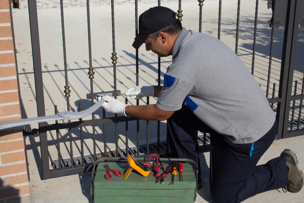 A person in a gray uniform kneels, fixing a metal gate with tools from a green toolbox on a sunny day.