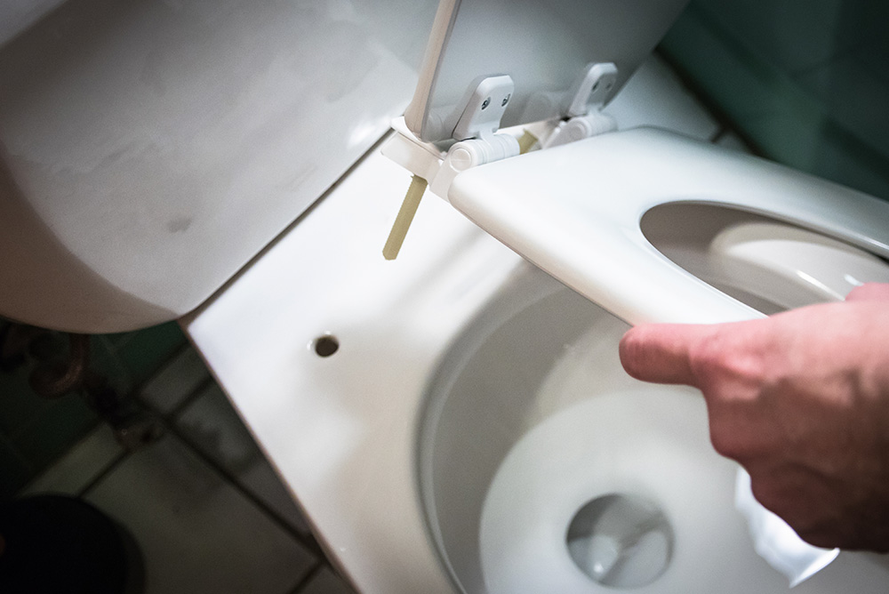A person’s hand is lifting a white toilet seat in a bathroom, revealing a clean, open toilet bowl beneath.
