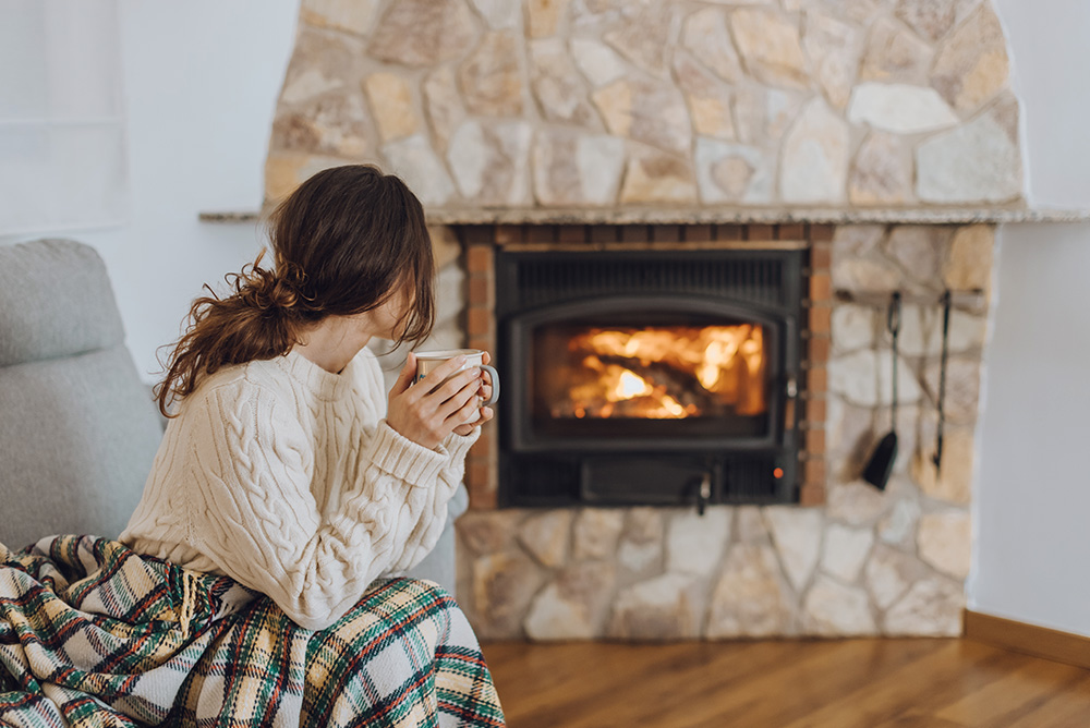 A woman in a cozy sweater drinks from a mug by a fireplace, wrapped in a plaid blanket, seated on a gray sofa.