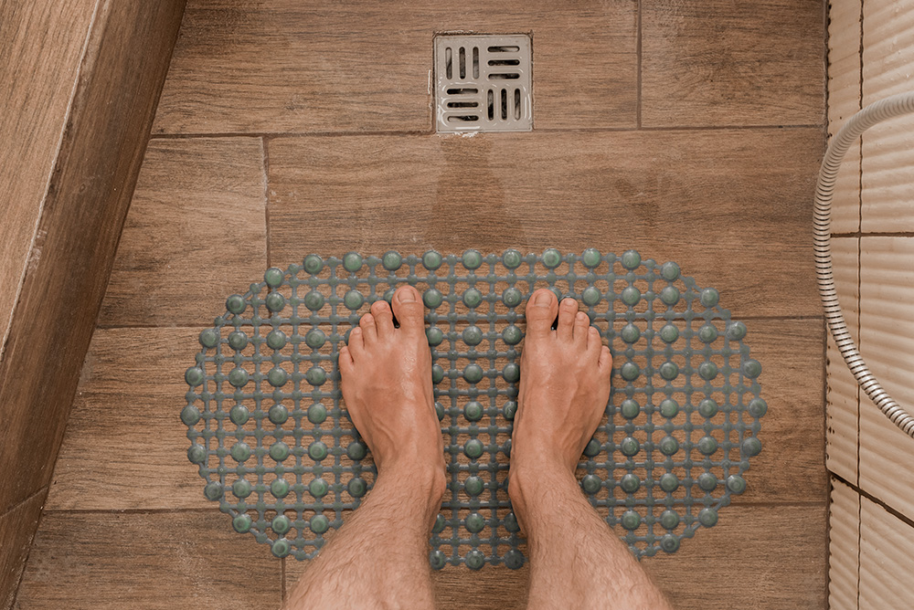 Two bare feet standing on a textured blue bath mat in a tiled shower with a metal drain.