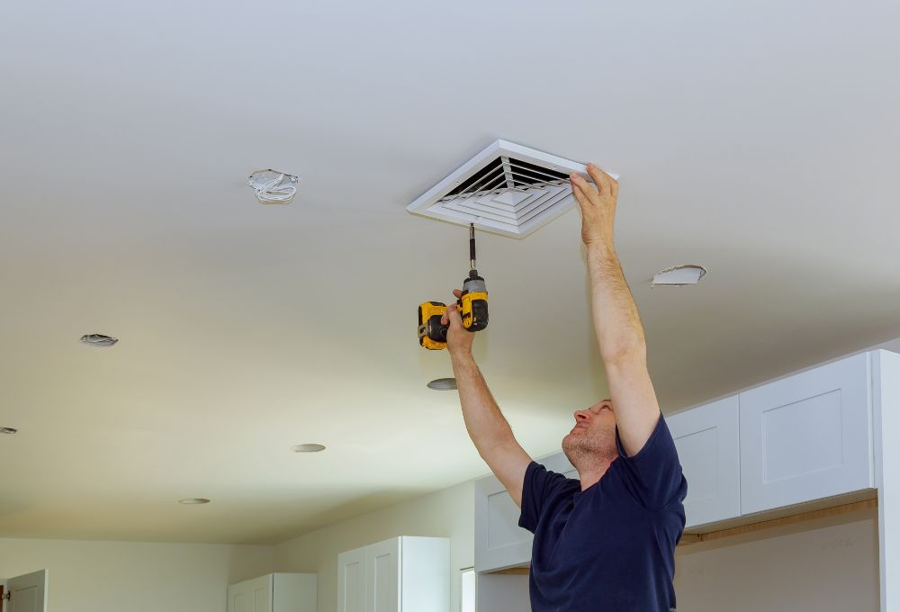 A man in a blue shirt uses a drill to install a ceiling air vent in a room with white cabinets.