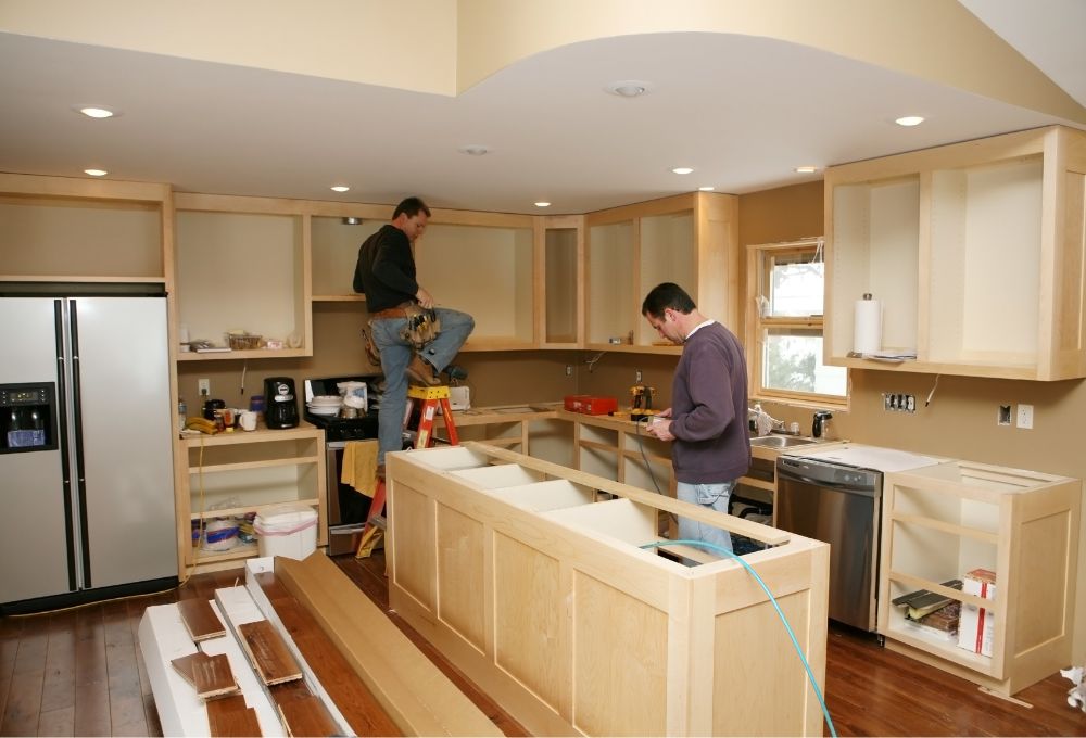 Two men work on installing wooden kitchen cabinets, one on a ladder and one checking a tool, in a partially finished kitchen.